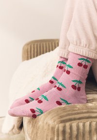 Vaaleanpunaiset sukat, joissa on kirsikkakuvio punaisine kirsikoineen ja vihreine lehtineen. Kangas näyttää pehmeältä ja tarjoaa mukavan istuvuuden.