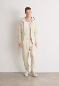 Crème-kleurige hoodie en bijpassende sweatpants van zachte stof, met een ontspannen pasvorm en een eenvoudig ontwerp, gecombineerd met lichte sneakers.