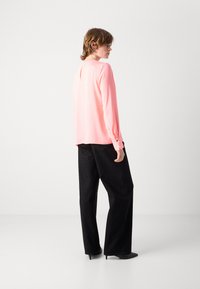Helle rosa Bluse mit langen Ärmel und einem leichten Rückenausschnitt, kombiniert mit lockeren schwarzen Hosen mit kleinen Glitzerpartikeln und Ankle Boots.