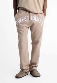 Pantalons de survêtement beiges avec une taille côtelée, "SWEET PANTS" imprimé en blanc sur le devant. Tissu doux avec une coupe ample.