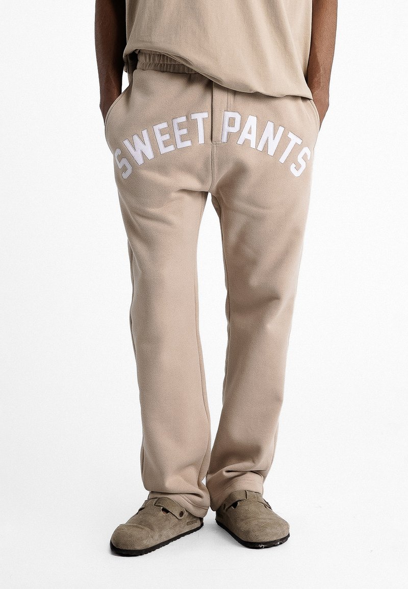 Pantalons de survêtement beiges avec une taille côtelée, "SWEET PANTS" imprimé en blanc sur le devant. Tissu doux avec une coupe ample.
