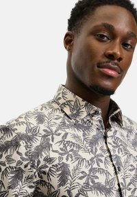 Junger Mann mit kurzen lockigen Haaren, der ein beige Knopfhemd mit schwarzen und grauen tropischen Blattmustern trägt und selbstbewusst in die Kamera schaut.