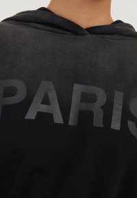 Sudadera negra con una textura suave, que presenta la palabra "PARÍS" en letras gruesas, brillantes y de color gris oscuro en el pecho y capucha con cordones.