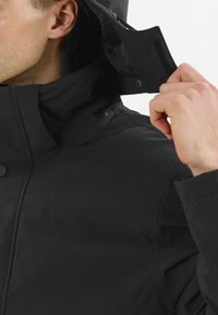 Svart vattentät jacka med hög krage, knappstängning och invändiga velcro-accenter; slät textur och minimal design.