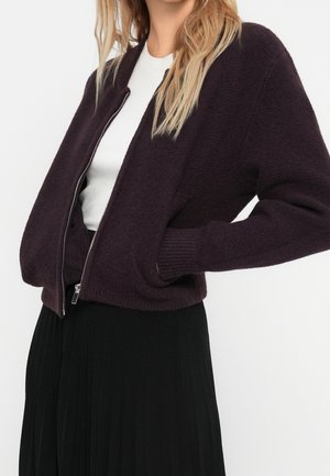 Femme portant un cardigan zippé violet foncé sur un haut blanc avec une jupe plissée noire, les mains dans les poches du cardigan.
