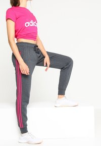 Różowa krótka koszulka z białym logo "adidas"; szare miękkie joggery z różowymi bocznymi paskami; model w białych butach sportowych.