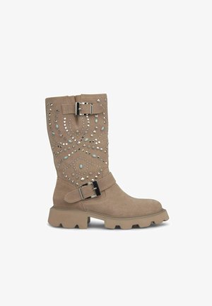 Kawowe zamszowe kozaki do kolan z dekoracyjnym wzorem w ćwieki i dwoma akcentami w postaci klamr. Gruba, wytrzymała podeszwa dopełnia całość.