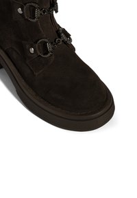 Brauner Veloursleder-Stiefelette mit rundlicher Spitze, mit dekorativen Riemen und Metallbeschlägen. Die Sohle ist strukturiert und leicht erhöht.