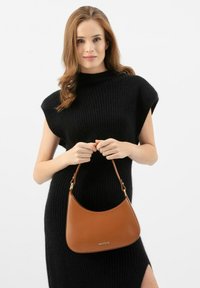 Sac à main en cuir marron avec un design incurvé, quincaillerie dorée et une sangle courte. Le modèle porte une robe en tricot noire. Apparence simple et épurée.