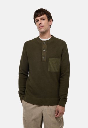 Pull en tricot vert olive avec un col rond, trois boutons, une finition texturée et une poche sur la poitrine. Porté avec un pantalon en velours côtelé beige.