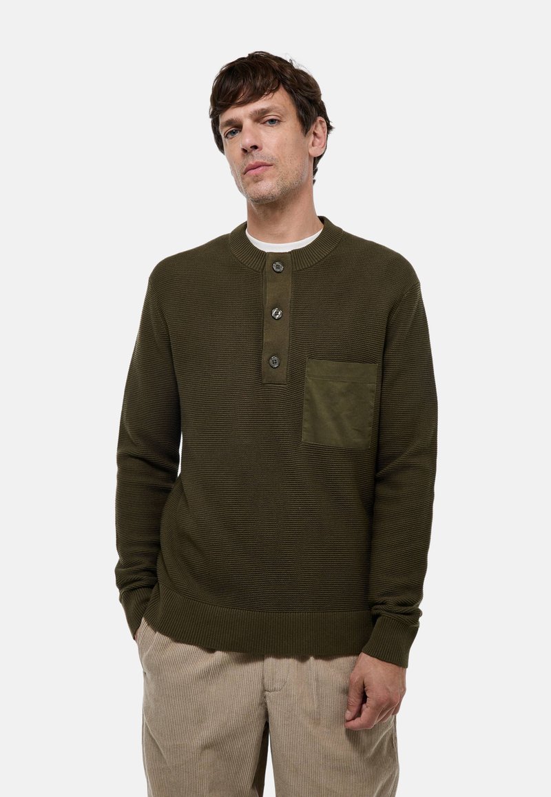 Pull en tricot vert olive avec un col rond, trois boutons, une finition texturée et une poche sur la poitrine. Porté avec un pantalon en velours côtelé beige.