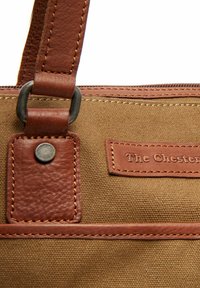 Sac en toile avec des accents en cuir marron, présentant une surface texturée, des coutures visibles, des accessoires métalliques et une étiquette de marque embossée sur le cuir.