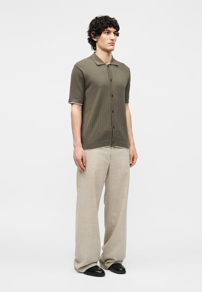 Άνδρας με σγουρά μαλλιά που φοράει μια ελιά κοντομάνικη πουκάμισο με κουμπιά, μπεζ φαρδιά παντελόνια και μαύρα παπούτσια, στέκεται μπροστά σε ένα μονόχρωμο φόντο.