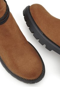 Rjavi antilopasti gležnjarji z teksturiranim zaključkom, ki imajo pleteno obrobo in črn gumijast podplat z globokim profilom za boljši oprijem in trajnost.