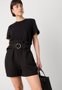 Haut noir à manches courtes associé à un short noir taille haute. La ceinture comporte un grand anneau métallique. Le modèle tient un sac à main noir texturé.