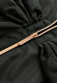 Svart pufferjacka med en rynkad textur. Har en roséguld dragkedja med detaljerad branding på dragfliken.