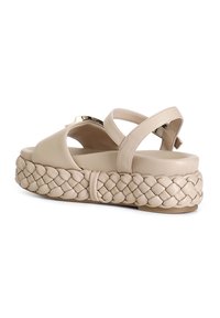 Beige leather sandal with braided platform sole, featuring an adjustable ankle strap and metallic accent. Smooth texture and rounded shape.