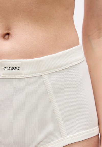 Κοντινό πλάνο στη μέση ενός ατόμου που φοράει λευκά ριμπ σορτς με μια μικρή ετικέτα «CLOSED» στη ζώνη μέσης.