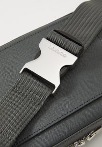 Cataramă metalică cu inscriptionarea "SANDRO", atașată de o curea din material texturat de culoare gri închis și o geantă cu fermoare vizibile.