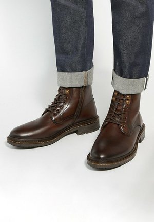 Brown leather ankle boots feature a rounded toe, lace-up design, and a textured rubber sole. Cuffs of dark denim jeans show a contrasting gray lining.