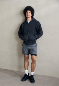 Čierna mikina na zips s prednými vreckami, šedé športové šortky, biele ponožky po členky a čierne tenisky. Model stojí pred neutrálnou stenou.