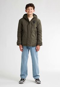 Jachetă căptușită verde măsliniu cu glugă, închidere cu fermoar și buzunare frontale, combinată cu blugi bleu deschis și adidași negri.