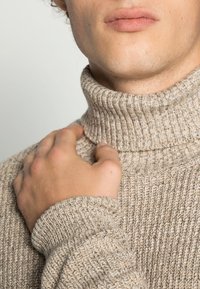 Beżowy sweter z prążkowanej dzianiny i golfem, z charakterystycznym wysokim kołnierzem i teksturowanym materiałem. Zbliżenie ręki opierającej się na okolicy ramienia.