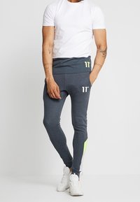 Grijze sportbroek met een aansluitend ontwerp, voorzien van een neongroene accent bij de taille en logodetails. Gekleed met een wit T-shirt.