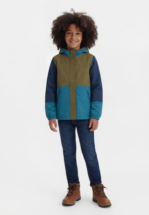 Lächendes Kind mit lockigem Haar, das eine blau-olivgrüne Kapuzenjacke, dunkle Jeans und braune Schnürstiefel trägt, vor weißem Hintergrund stehend.