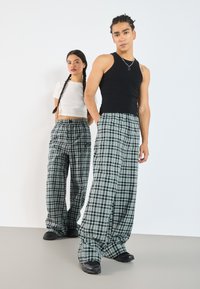 Schwarzes Tanktop und weißes Croptop kombiniert mit karierten, weiten Hosen in Grün und Schwarz, die einen elastischen Bund und Taschen haben.