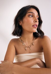 Gold chain necklace featuring irregular links and fish-shaped pendants. Model wearing strapless, neutral-colored top. Smooth skin texture.