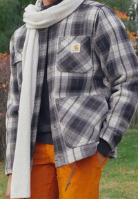 Grau und schwarz karierter Flanellhemdjacke mit zwei Brusttaschen, einem Logo-Patch und einem cremefarbenen Strickschal, kombiniert mit orangefarbenen Cordhosen.