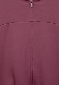 Detailný záber na fialovú látku s viditeľným centrálnym zipsom a horizontálnym švom cez oblasť hrudníka.