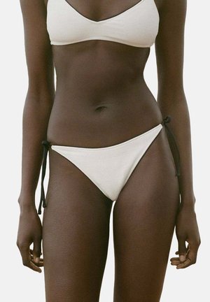 Bikini deux pièces en tissu blanc. Le haut a une forme triangulaire, tandis que le bas est taille basse avec des liens ajustables noirs sur les côtés.