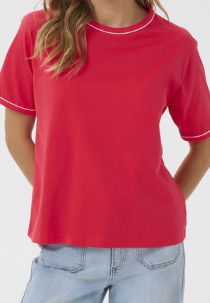 Kvinde iført en rød T-shirt med korte ærmer og hvide kantbånd på krave og ærmer, kombineret med lyseblå cowboybukser.