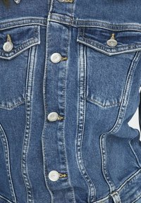 Vue de face d'une veste en jean bleue avec des boutons argentés et deux poches poitrine à rabats boutonnés, portée par une personne.