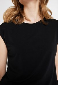 Femme aux cheveux châtain clair mi-longs portant un haut noir sans manches uni sur un fond neutre.