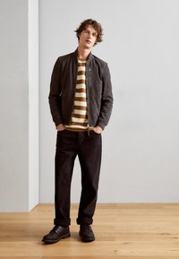 Chaqueta bomber negra de ante, camiseta de manga larga a rayas amarilla y blanca, pantalones oscuros y botas marrones con cordones están de pie sobre un suelo de madera.