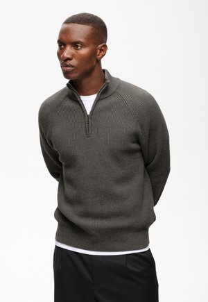 Homme portant un pull côtelé gris foncé à demi-fermeture éclair sur une chemise blanche avec un pantalon noir, debout les mains dans le dos.