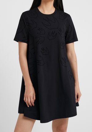 Mujer que lleva un vestido negro holgado de manga corta con un sutil patrón de siluetas de pájaros en la parte superior delantera y en las mangas.