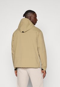 Beige hooded jacket made of lightweight fabric, featuring a relaxed fit, zippered pockets, and elastic cuffs. Visible stitching details.