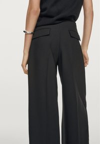 Person iført sorte brede bukser med baglommer og en ærmeløs sort top, sølvarmbånd på venstre håndled, stående.