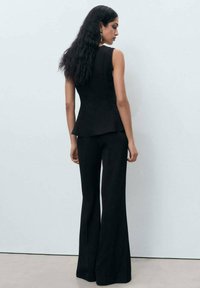 Černý bezrukávový peplum top s upnutým pasem, spárovaný s kalhotami volného střihu. Hladká textura a rafinovaný design, pohled zezadu.