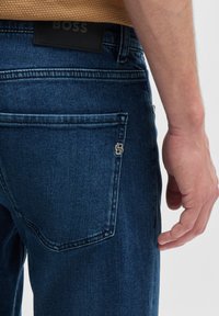 Dark blue denim jeans featuring a silver logo accent on the back pocket, reinforced stitching, and a textured black leather patch on the waistband.