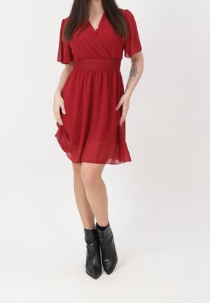 Mujer con vestido rojo hasta la rodilla con mangas fluidas y botas negras hasta el tobillo, de pie con las manos cerca de las caderas sobre un fondo blanco.