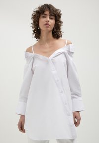 Chemise blanche, décolletée et à manches longues, dotée d'une fermeture à boutons sur le devant et d'un léger éclat dans le tissu. Coupe droite avec une coupe décontractée.