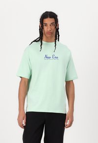 Mintgrønne bomulds-T-shirt med korte ærmer, der har et blåt "New Era"-logo trykt på brystet, parret med sorte bukser.
