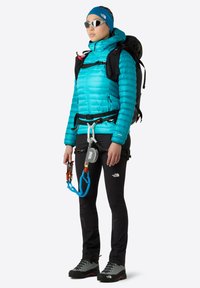 Klart blått foderjacka i kombination med svarta klätterbyxor och robusta kängor. En sele med utrustning och solglasögon fulländar den utomhuslook.