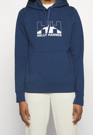 Osoba nosící modrou mikinu Helly Hansen s bílým logem a textem, v kombinaci s světlými kalhotami, zobrazená od krku po pás.