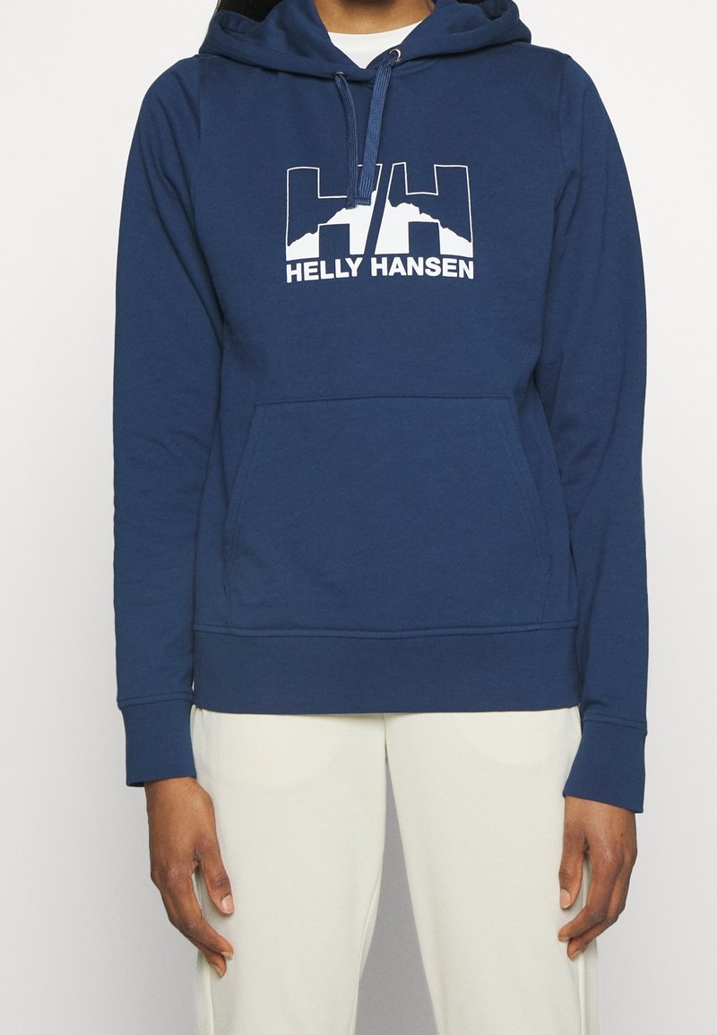 Personne portant un sweat à capuche Helly Hansen bleu avec un logo et un texte blancs, associé à un pantalon clair, visible du cou à la taille.